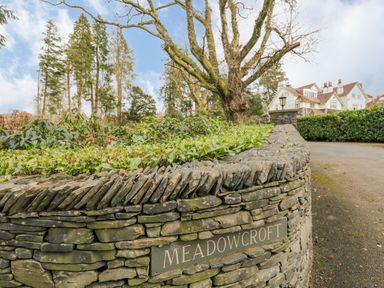 A stone wall with plants on top and a sign reading Meadowcroft with a large tree and a house in the background at Lake House Loft in Bowness-On-Windermere