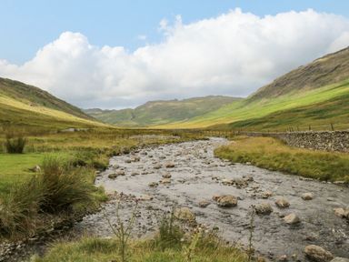 A river flowing through a valley with grassy hills and a stone wall at Lake House Loft in Bowness-On-Windermere