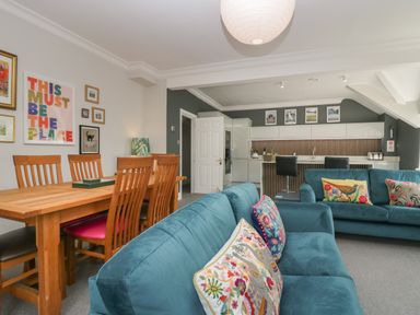 An open plan living room with blue sofas and colorful cushions a wooden dining table with chairs and a kitchen area with bar stools at Lake House Loft in Bowness-On-Windermere