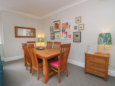 A dining room with a wooden table and six chairs a side table with a lamp and framed pictures on the wall at Lake House Loft in Bowness-On-Windermere