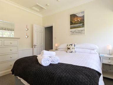 A bedroom with a bed with white linens and a black blanket rolled towels on the bed a chest of drawers with mirror a door and a framed poster on the wall at Lake House Loft in Bowness-On-Windermere