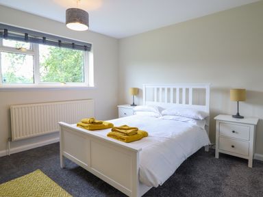 A bedroom with a white bed frame white bedding yellow towels on the bed two white bedside tables with yellow lamps a window and grey carpet at The Wardens House in Tregynon near Newtown