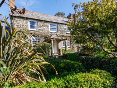 A stone cottage with white framed windows and a garden with bushes and trees at Aarons Cottage in St Teath