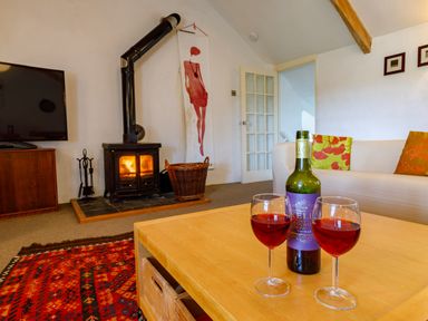 A living room with a wood stove a television a wooden table with two glasses of red wine and colorful cushions on a white sofa at Aarons Cottage in St Teath