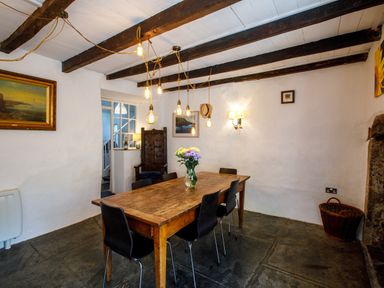 A dining room with a wooden table and six chairs a vase with flowers hanging light bulbs exposed wooden ceiling beams and a wicker basket at Aarons Cottage in St Teath