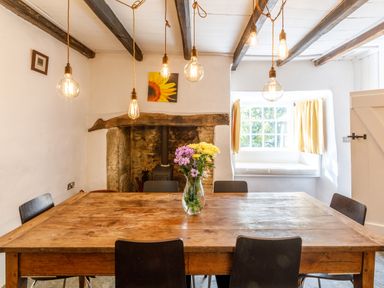 A dining room with a wooden table flowers in a vase hanging light bulbs and a window seat at Aarons Cottage in St Teath