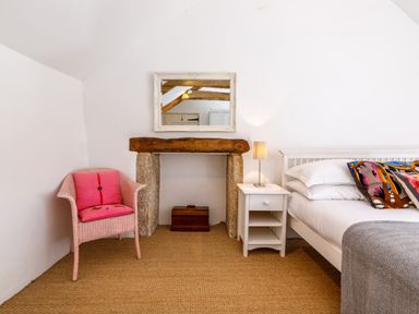 A bedroom with a bed, bedside table with lamp, pink chair with cushion, and a fireplace with mirror at Aarons Cottage in St Teath