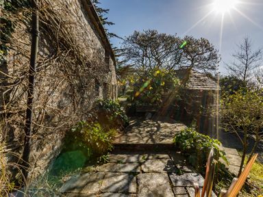 An outdoor stone patio with plants and trees next to a stone building under bright sunlight at Aarons Cottage in St Teath