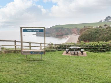 A green grassy area with a wooden sign #ladrambay and a picnic table overlooking a beach and cliffs at The Retreat in Otterton