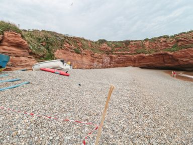 A pebble beach with a white boat and a red kayak near red rock cliffs with two people at the water edge at The Retreat in Otterton