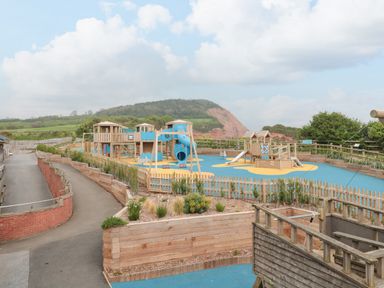 An outdoor playground with slides swings and climbing structures on a blue and yellow surface with hills in the background at The Retreat in Otterton