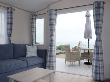 A living room with a blue sofa and wooden coffee table next to a sliding door opening to a deck with wicker chairs and a table with a bottle and glasses at The Retreat in Otterton