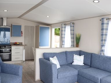 A living room with blue sectional sofa two pillows blue plaid curtains partition wall and kitchen with blue cabinets at The Retreat in Otterton