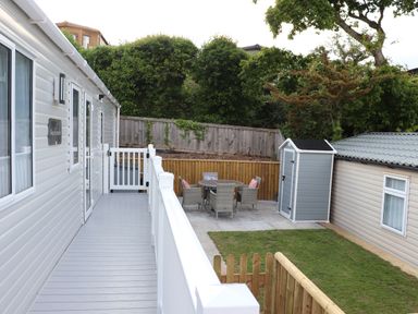 An outdoor patio area with a round table and chairs next to a small shed and grassy lawn at The Retreat in Otterton