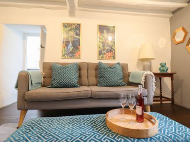 A living room with a brown sofa with teal patterned pillows and blankets a wooden side table with a lamp and decorative item two framed pictures and a wooden tray on a patterned teal ottoman with a wine bottle and two glasses at 3 Ivy Court in Dolgellau