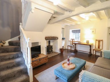 A living room with a staircase a wood stove a wooden table and chairs a TV and a patterned ottoman at 3 Ivy Court in Dolgellau