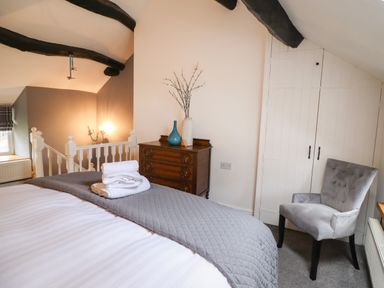 A bedroom with a bed folded towels a wooden dresser with vases a grey upholstered chair and white built in cupboards at 3 Ivy Court in Dolgellau