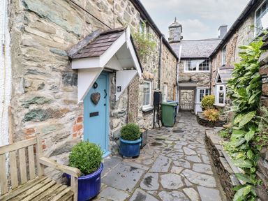 An outdoor stone pathway between stone buildings with a blue door bench green plants and bins at 3 Ivy Court in Dolgellau