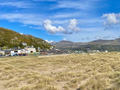 A sandy beach with grass and hills in the background with buildings near the shoreline at Deu-Ddrws in Barmouth