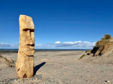 A wooden sculpture carved like a face standing on a sandy beach with dunes and the sea in the background at Deu-Ddrws in Barmouth