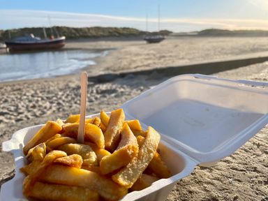 A close up of chips in a takeaway box on a beach with boats in the water at Deu-Ddrws in Barmouth
