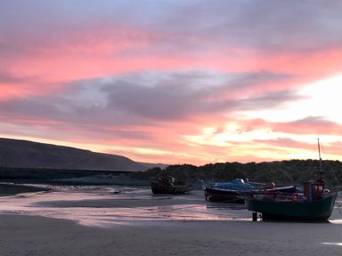 Boats resting on a sandy shore with hills and a pink and purple sunset sky at Deu-Ddrws in Barmouth