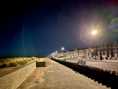 A nighttime view of a seaside promenade with street lamps and benches along a row of buildings at Deu-Ddrws in Barmouth