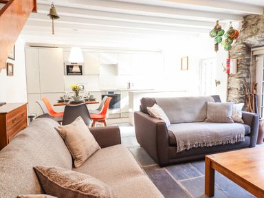 An open living room and kitchen with two sofas a wooden dining table with chairs and white kitchen cabinets at Deu-Ddrws Barmouth