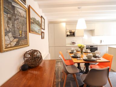 A dining area with a wooden table set with dishes and glasses surrounded by gray and orange chairs and paintings on the wall in a kitchen at Deu-Ddrws in Barmouth