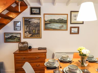 A dining area with a wooden table set with plates bowls glasses and cutlery a wooden chest of drawers with decorations and framed pictures on the wall at Deu-Ddrws in Barmouth