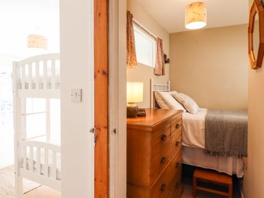 A bedroom with a wooden chest of drawers, a bed with pillows and a blanket, a small window with curtains, and a white bunk bed visible through an open doorway at Deu-Ddrws in Barmouth