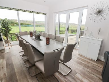 A dining room with a long table surrounded by chairs next to large windows and glass doors at Ty Marlyn in Crofty
