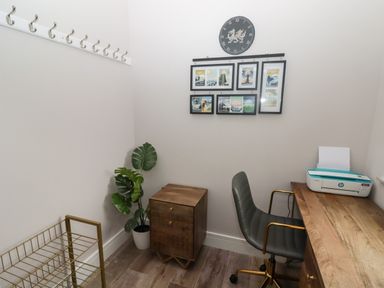 A small office with a wooden desk a gray office chair a wooden cabinet a plant a wall clock and framed pictures at Ty Marlyn in Crofty