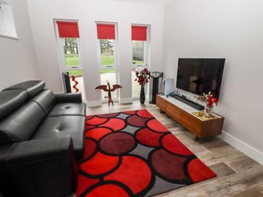A living room with a black leather sofa a red and black patterned rug a wooden TV stand with a flat screen TV and three tall windows with red blinds at Ty Marlyn in Crofty