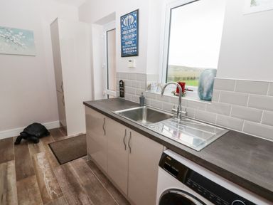 A kitchen sink area with cabinets washing machine and a window showing outdoor view at Ty Marlyn in Crofty