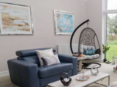 A living room with a blue armchair, hanging chair, side table, two paintings on wall, and decorative items on coffee table at Ty Marlyn in Crofty