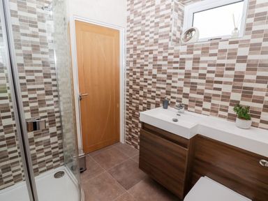 A bathroom with a wooden door a corner shower unit a white sink with wooden cabinetry and a small window with decorative items at Ty Marlyn in Crofty