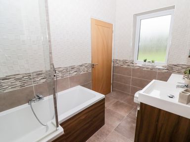 A bathroom with a bathtub shower, wooden door, window with plants on the sill and a sink with a vanity unit at Ty Marlyn in Crofty