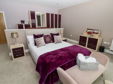 A bedroom with a double bed pillows and a purple blanket a bench with a basket of towels two bedside tables with lamps and a dresser with a mirror at Ty Marlyn in Crofty