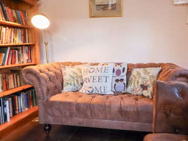 A brown couch with decorative pillows near a wooden bookshelf and a floor lamp at Rhubarb Cottage in Ufford near Woodbridge