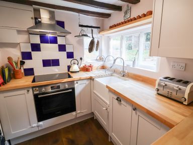 A kitchen with wooden countertops white cabinets a black oven a stove with a stainless steel hood a sink under a window and utensils at Rhubarb Cottage in Ufford near Woodbridge