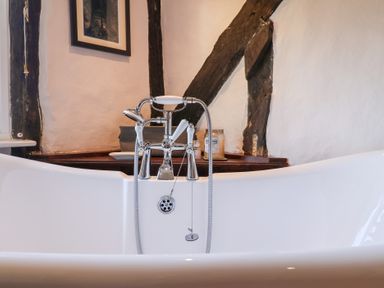 A close up of a white freestanding bathtub with chrome taps and a handheld shower in a bathroom with wooden beams at Rhubarb Cottage in Ufford near Woodbridge