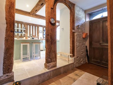 An entrance area with wooden door and brick wall opening into a kitchen with wooden beams and stools at a counter at The Bridewell in Woodbridge