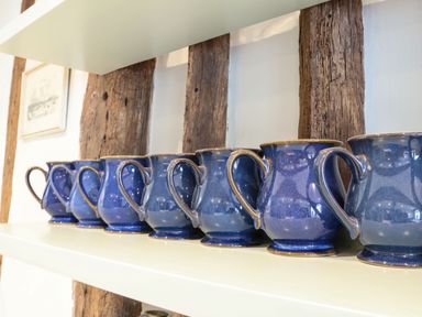 A row of blue ceramic mugs on a white shelf with wooden beams on the wall at The Bridewell in Woodbridge