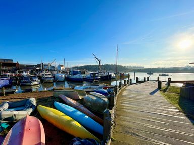 A marina with boats docked near a curved wooden walkway and kayaks on the shore at The Bridewell in Woodbridge