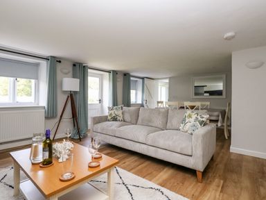 A living room with a beige sofa two floral cushions a wooden coffee table with glasses and a bottle a floor lamp and a dining table with chairs at Dairy Cottage in Abbotsbury