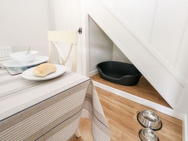 A dining table with a plate of bread, a bowl, and cutlery next to a chair and a pet bed under a staircase with two pet bowls on the floor in Dairy Cottage in Abbotsbury