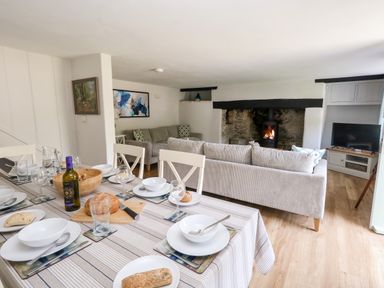 A dining table set with plates glasses and bread next to a living area with sofas a fireplace and a television at Dairy Cottage in Abbotsbury