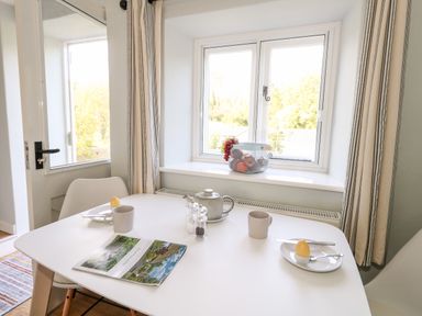 A dining area with a white table set with cups plates boiled eggs a teapot and a brochure by windows with curtains at Dairy Cottage in Abbotsbury