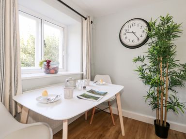 A dining area with a white table set for two near a window and a potted plant with a clock on the wall at Dairy Cottage in Abbotsbury